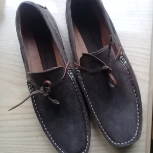 Peut inclure: Une paire de mocassins en daim marron avec des surpiq&ucirc;res blanches autour des bords. Les mocassins ont des lacets en cuir marron et sont nou&eacute;s en un n&oelig;ud.