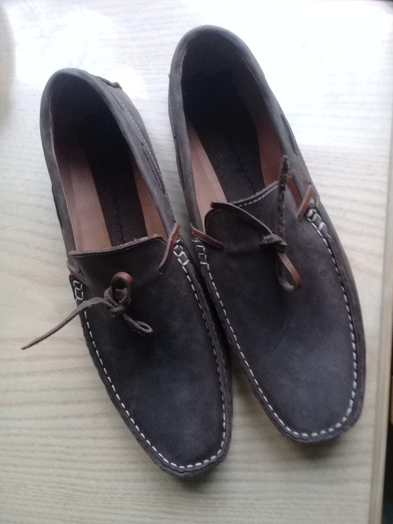 Peut inclure: Une paire de mocassins en daim marron avec des surpiq&ucirc;res blanches autour des bords. Les mocassins ont des lacets en cuir marron et sont nou&eacute;s en un n&oelig;ud.
