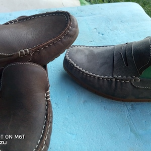 Peut inclure: Deux paires de mocassins en cuir. Une paire est marron avec des coutures blanches et l'autre paire est grise avec des coutures blanches. Les mocassins sont sur une surface bleue.