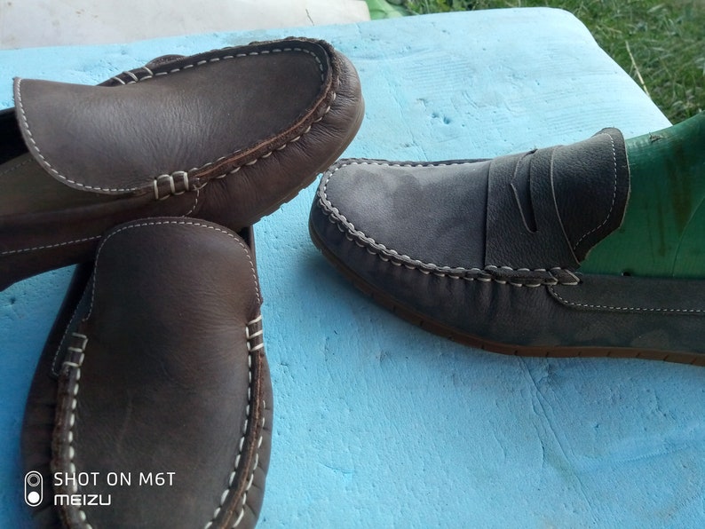 Peut inclure: Deux paires de mocassins en cuir. Une paire est marron avec des coutures blanches et l'autre paire est grise avec des coutures blanches. Les mocassins sont sur une surface bleue.
