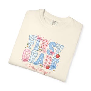 Może przedstawiać: Kremowy t-shirt z nadrukiem graficznym przedstawiającym napis "FIRST GRADE" dużymi, wzorzystymi literami blokowymi. Litery ozdobione są niebiesko-różową kratką, motywami kwiatowymi i truskawkami, z akcentami w postaci małych różowych kokardek i stokrotek.