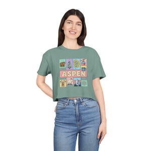 May include: A sage green cropped t-shirt featuring a central graphic with the text "ASPEN" surrounded by small illustrations of a bear, pine tree, yellow leaves, ski gondola, hot cocoa, skis, a mountain goat, mountains, and a snowflake.