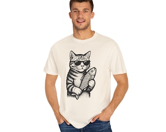 May include: Cream-coloured t-shirt with a black and white illustration of a cat wearing sunglasses and holding a fish. The cat has striped fur. The t-shirt is a casual style.
