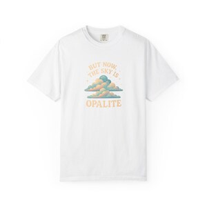 Pode incluir: Camiseta branca com um gráfico de nuvens em tons pastel e o texto "BUT NOW THE SKY IS OPALITE". O design utiliza uma paleta de cores azul claro, pêssego e creme. Gola redonda e mangas curtas.