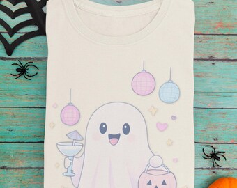 Puede incluir: Camiseta color crema con un fantasma de dibujos animados sosteniendo un cóctel y un cubo de calabaza. El fantasma está rodeado de adornos de bolas de discoteca, estrellas y corazones. Ropa temática de Halloween.