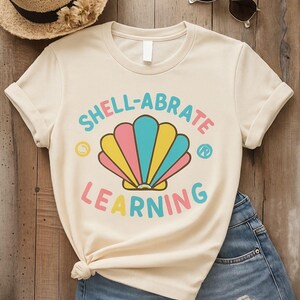 May include: Cream-colored t-shirt with the words "SHELL-ABRATE LEARNING" in colorful text and a seashell graphic. The shirt is paired with denim shorts, a straw hat, and sunglasses, suggesting a summer or beach theme.