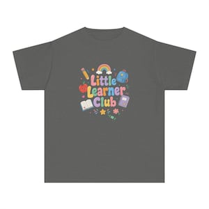Op de afbeelding: Een donkergrijs t-shirt met korte mouwen en een kleurrijke grafische print met de tekst Little Learner Club. Het ontwerp bevat een regenboog, een potlood, een rode appel, een blauwe rugzak, een open boek, een notitieboek, een ster, een bloem en een groen krijtje.