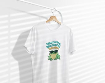 May include: White t-shirt with a graphic of a green frog wearing sunglasses. The text above the frog reads "UNBOTHERED, MOISTURIZED, THRIVING" in teal and yellow. The shirt is hanging on a wooden hanger.