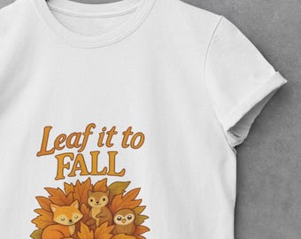 Könnte beinhalten: Weißes T-Shirt mit dem orangefarbenen Schriftzug "Leaf it to FALL". Darunter eine Grafik mit einem Fuchs, einem Eichhörnchen und einer Eule in Herbstblättern. Das T-Shirt hängt an einem Holzbügel.