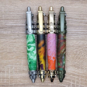 Puede incluir: Cuatro bolígrafos tácticos con diferentes patrones de espiral de colores y detalles metálicos. Los bolígrafos están dispuestos en una fila de izquierda a derecha: plata, oro, oro y verde.