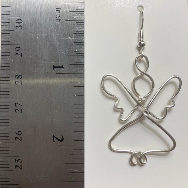 Puede incluir: Un pendiente de &aacute;ngel de alambre plateado. El dise&ntilde;o del &aacute;ngel presenta una cabeza, alas y una parte inferior en forma de falda. El pendiente mide aproximadamente 5 cm de largo, medido por la regla en pulgadas de la imagen.