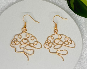 May include: A pair of gold-toned wire earrings shaped like a human brain. Each earring features a continuous, swirling metal line that forms the anatomical structure of a brain, suspended from a simple gold-toned hook ear wire for pierced ears.