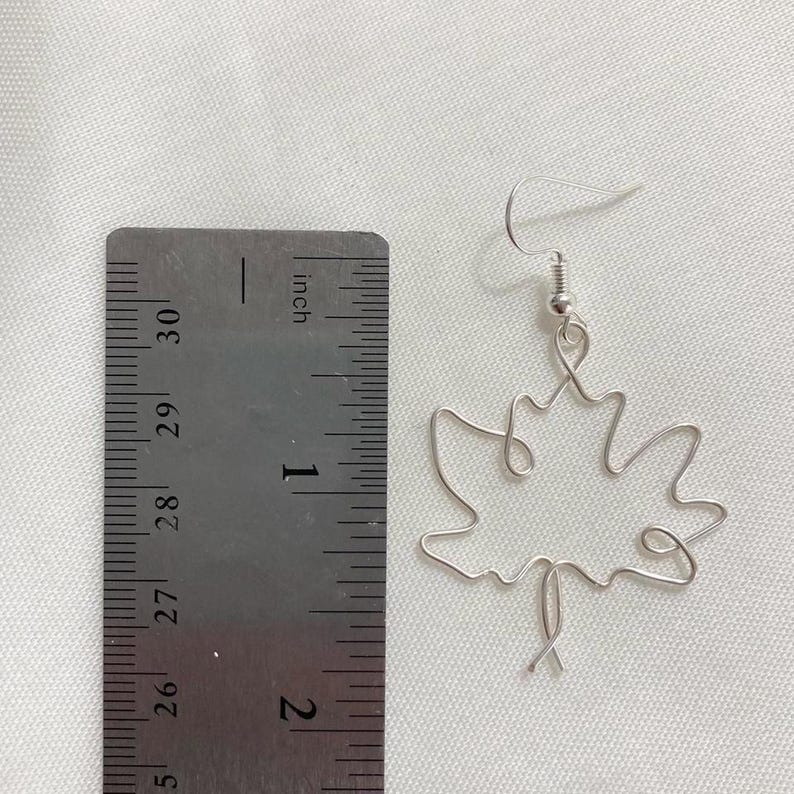 May include: Silver wire earrings shaped like a maple leaf. The earrings are approximately 2.5 cm long and are shown next to a ruler.