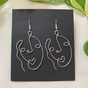 May include: A pair of silver wire earrings shaped like abstract faces. Each earring features a simple line drawing of a face with a heart shape in the center.