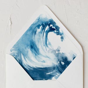 Puede incluir: Un sobre blanco con un diseño de acuarela de una ola azul en el interior. El diseño de la ola presenta tonos de azul y blanco, creando un efecto dinámico y artístico. El sobre está abierto, revelando la obra de arte.