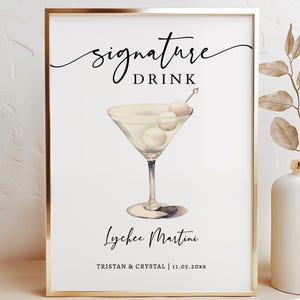 May include: A rectangular sign in a gold frame featuring the text signature DRINK, an illustration of a martini glass with three lychees on a pick, the text Lychee Martini, and the names TRISTAN & CRYSTAL with the date 11.05.20xx below.