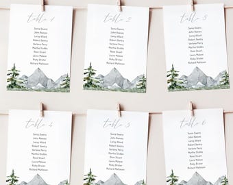 May include: Six white table cards with watercolor mountain and tree illustrations, suspended by clothespins. Each card features a table number and guest names in elegant script, perfect for a wedding or event.