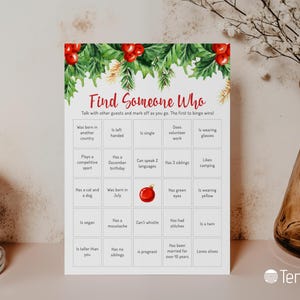 May include: A festive Christmas-themed bingo game card with a watercolor design. The card features a green and red holly border and the text "Find Someone Who." The bingo squares contain prompts like "Is vegan" and "Has a mustache."