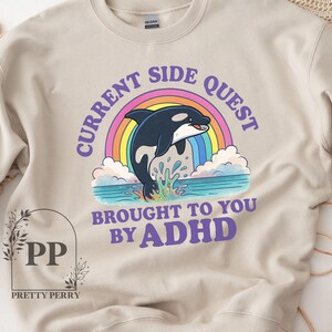 May include: A beige sweatshirt with a graphic of a black and white orca jumping over a rainbow. The text "Current Side Quest Brought To You By ADHD" is printed in purple on the sweatshirt.
