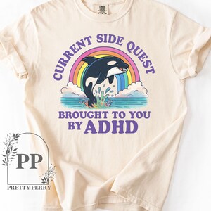 May include: A beige t-shirt with a graphic of a rainbow and an orca whale jumping out of the water. The text "Current Side Quest Brought To You By ADHD" is printed in purple on the shirt.