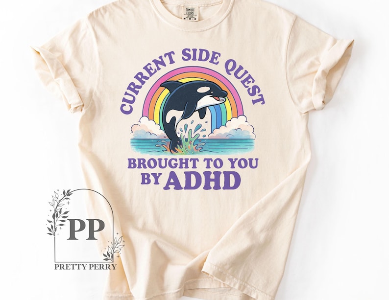 May include: A beige t-shirt with a graphic of a rainbow and an orca whale jumping out of the water. The text "Current Side Quest Brought To You By ADHD" is printed in purple on the shirt.