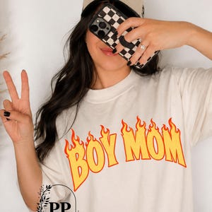 May include: A beige t-shirt featuring the text BOY MOM in yellow letters with orange flame outlines. A beige trucker hat with the text THE MOM CLUB COLLECTIVE in black script is worn. A black and white checkered phone case is held in front of the face.