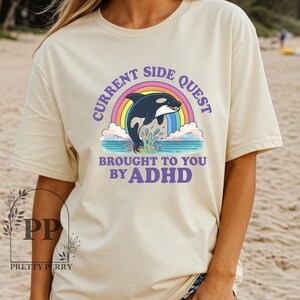 May include: A beige t-shirt with a graphic of a black orca jumping out of the water with a rainbow behind it. The text "Current Side Quest Brought to You by ADHD" is printed on the shirt.