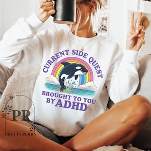 May include: White sweatshirt with a rainbow and an orca graphic. The text reads "Current Side Quest Brought To You By ADHD".