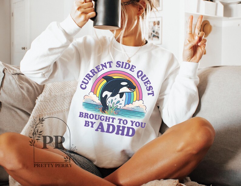 May include: White sweatshirt with a rainbow and an orca graphic. The text reads "Current Side Quest Brought To You By ADHD".