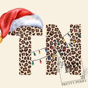 Puede incluir: Las letras "TN" están decoradas con un estampado de leopardo y una cadena de luces navideñas de colores. La letra "T" tiene un gorro de Papá Noel rojo con una campana dorada.