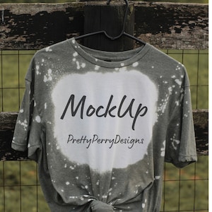 Może przedstawiać: Zielona koszulka z białymi plamami wybielacza i tekstem "MockUp PrettyPerryDesigns" z przodu. Koszulka wisi na drewnianym płocie.