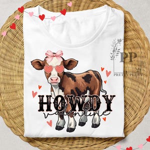 Pu&ograve; includere: Maglietta bianca con una mucca marrone e bianca che indossa occhiali da sole a forma di cuore rosa e un fiocco rosa. Il testo "Howdy Valentine" &egrave; stampato in nero sulla maglietta.