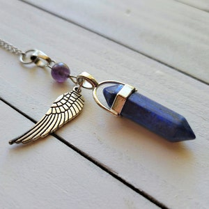 Könnte beinhalten: Eine silberne Kettenkette mit einem blauen Lapislazuli-Kristall-Anhänger, einem silbernen Flügel-Anhänger und einer kleinen violetten Amethyst-Perle.