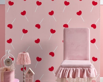 May include: A pink chair with a ruffled skirt and a white base sits in front of a pink wall with a repeating pattern of red heart-shaped lollipops.  Two pink boxes with white trim are on the left side of the image, along with a silver lamp with a pink lampshade and a small white house figurine.
