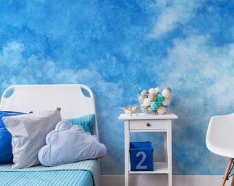 Puede incluir: Una cama blanca con ropa de cama y almohadas azules en una habitación con un mural de pared nublado azul y blanco. También hay una mesita de noche blanca con un cajón y una silla blanca en la habitación.