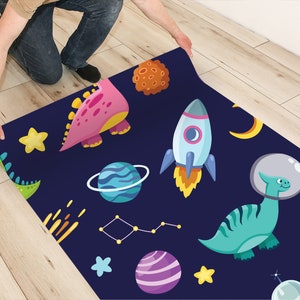 May include: A blue and black space-themed rug with dinosaurs, planets, stars, and rockets. The rug is being rolled up by a person's hand.