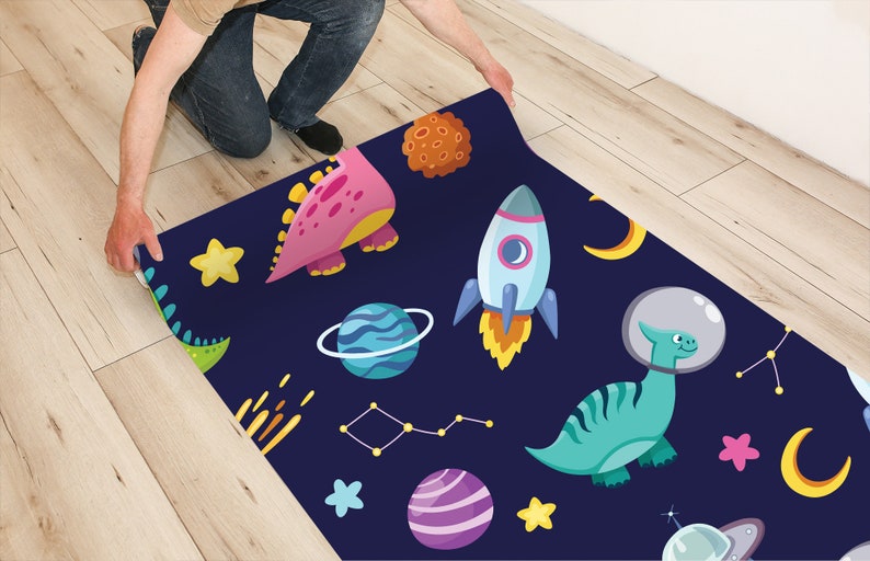 May include: A blue and black space-themed rug with dinosaurs, planets, stars, and rockets. The rug is being rolled up by a person's hand.