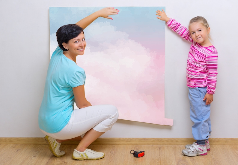 May include: A woman and a girl are putting up a large pink and blue watercolor wall mural. The mural features a cloudy sky design.