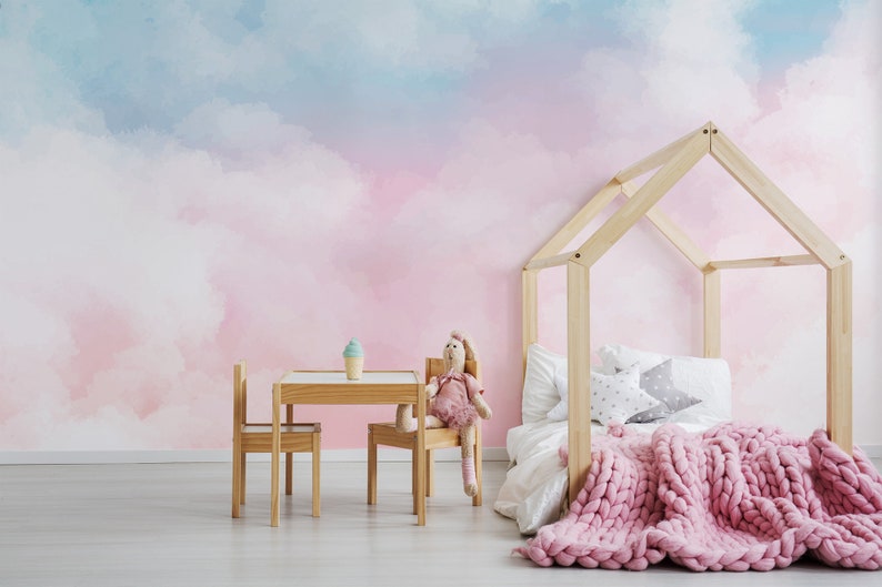 May include: A pink and blue watercolor cloud wallpaper with a wooden house bed frame, a pink knitted blanket, and a small wooden table and chairs.