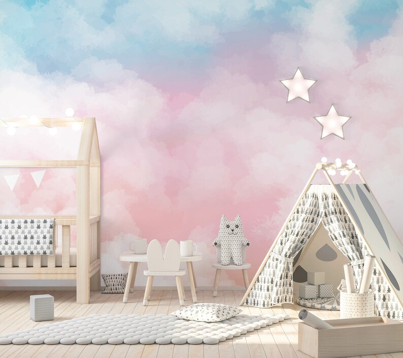 May include: A pink and blue watercolor cloud nursery wall mural with a white crib, a white teepee tent, a white bunny chair, and a white stuffed animal.