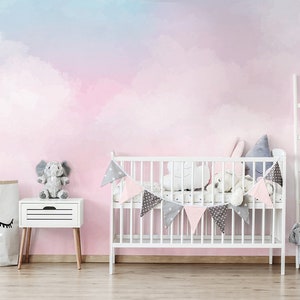 May include: A nursery room with a pink and blue watercolor cloud wall mural. The room features a white crib, a gray armchair, a wooden table and chairs, and a wooden shelf with a baby mobile.