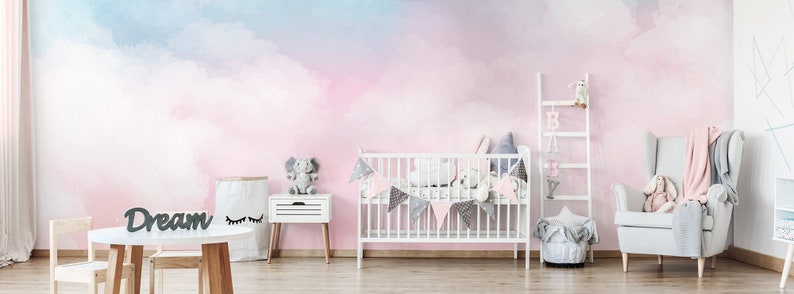May include: A nursery room with a pink and blue watercolor cloud wall mural. The room features a white crib, a gray armchair, a wooden table and chairs, and a wooden shelf with a baby mobile.