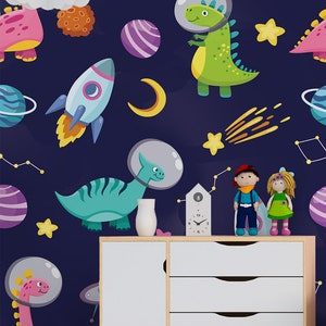 May include: A dark blue wallpaper with a space theme featuring dinosaurs wearing astronaut helmets, rockets, planets, stars, and a crescent moon. The wallpaper is displayed in a room with a white dresser, a white vase, and a set of colorful wooden blocks.