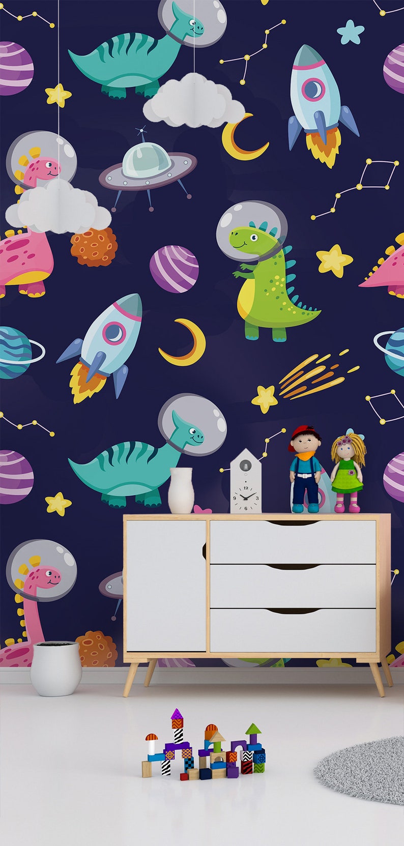 May include: A dark blue wallpaper with a space theme featuring dinosaurs wearing astronaut helmets, rockets, planets, stars, and a crescent moon. The wallpaper is displayed in a room with a white dresser, a white vase, and a set of colorful wooden blocks.