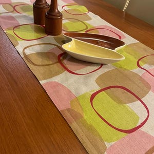 Può includere: Un runner da tavola con un motivo ripetitivo di quadrati arrotondati nei toni del rosa, verde lime e marrone. Due macina sale e pepe cilindrici in legno sono posizionati in verticale, accompagnati da un piatto in ceramica a due sezioni a forma di rene.