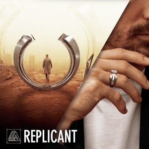 May include: A silver ring with a geometric design is shown on a person's finger. The ring has a unique shape with a central stone. The word "REPLICANT" is displayed in white text at the bottom of the image.