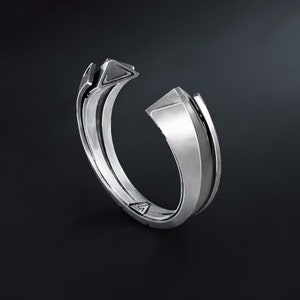 May include: A silver-colored ring with a geometric design. The ring features a split band with angular, raised elements and a triangular accent. The ring is set against a dark background.