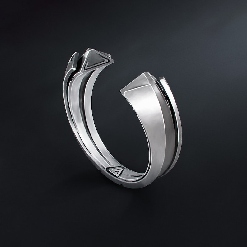 May include: A silver-colored ring with a geometric design. The ring features a split band with angular, raised elements and a triangular accent. The ring is set against a dark background.