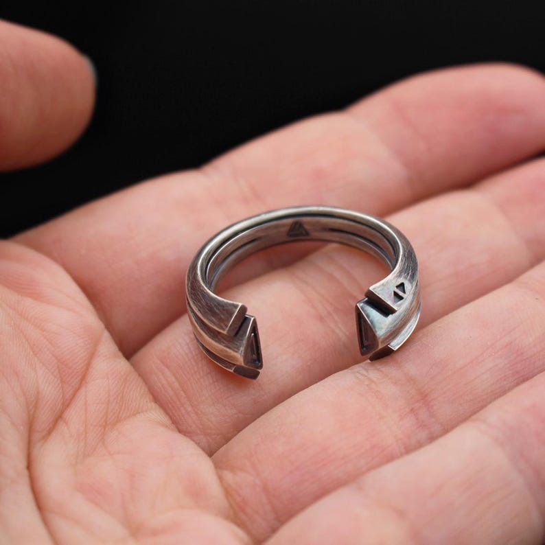 May include: A silver ring with a unique geometric design. The ring features a split band with angular cutouts and a small triangle detail. The ring is held in a hand against a dark background.