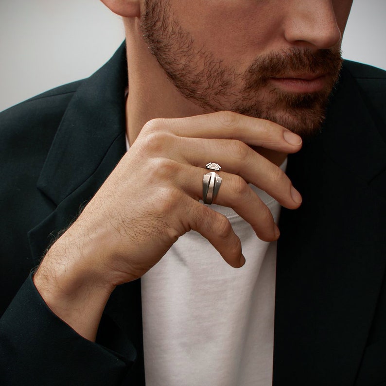 May include: A silver ring with a geometric design. The ring features a central, angular gemstone and a split band. The ring is worn on a finger, showcasing its modern and minimalist aesthetic. The ring's design is bold and eye-catching.